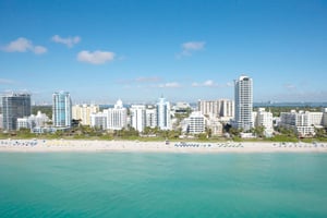 A wide-angle, high-altitude shot of a bustling beachfront. The foreground shows shimmering turquoise ocean water meeting a strip of white sand dotted with tiny figures and clusters of blue and yellow beach umbrellas. A dense line of white and light-blue modern skyscrapers and hotels runs parallel to the shore, with a clear blue sky and a few light clouds in the background